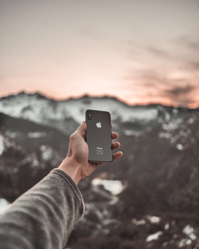 A person holds up a smartphone with snowy mountains in the background during sunset.