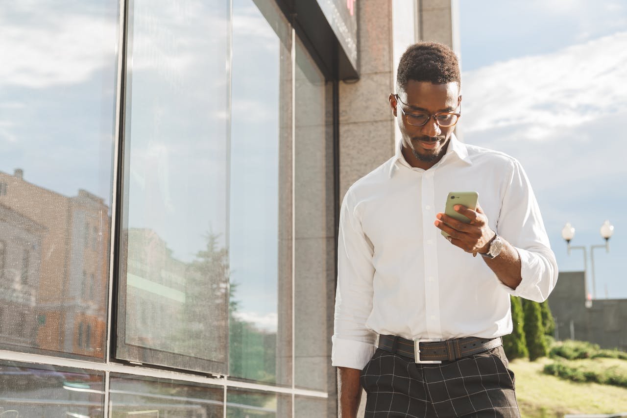 Confident young professional in eyeglasses checks phone messages while walking outside modern building.